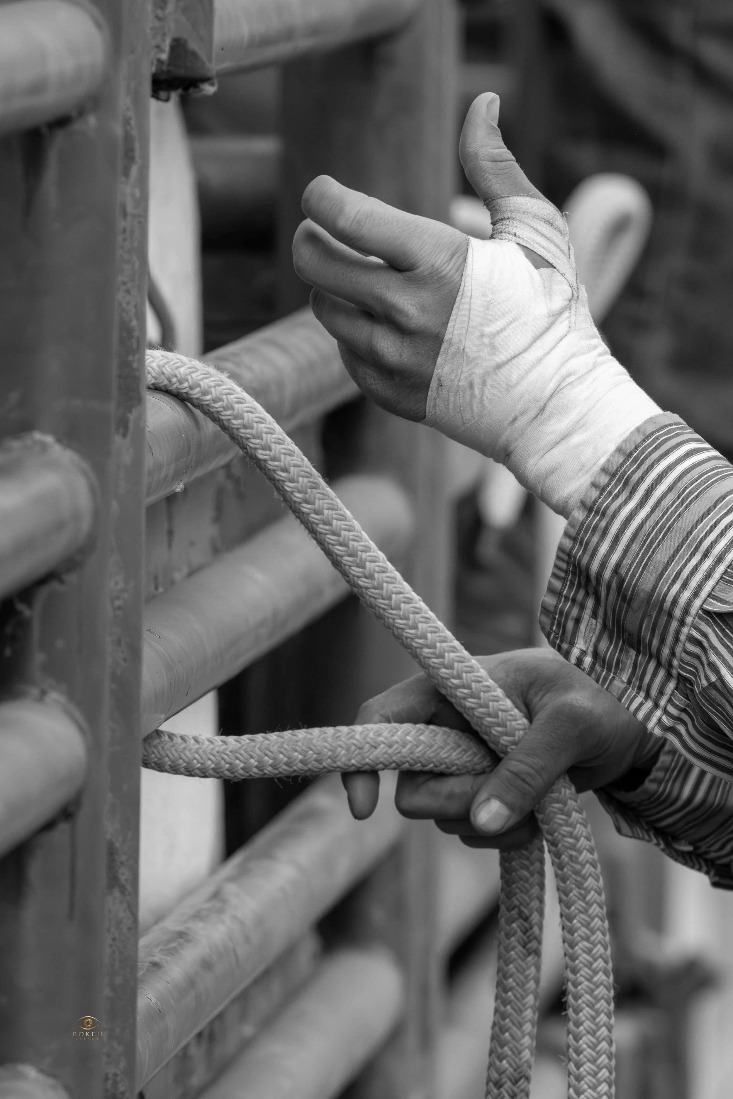 Close-up of a person's hand with a bandaged thumb, gripping a rope and a bamboo railing in a black-and-white photo.