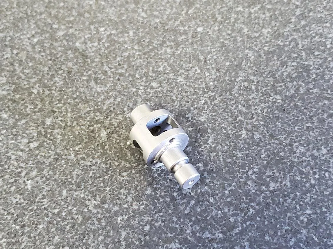 A small metallic mechanical part lying on a gray textured surface, possibly made of metal and plastic.