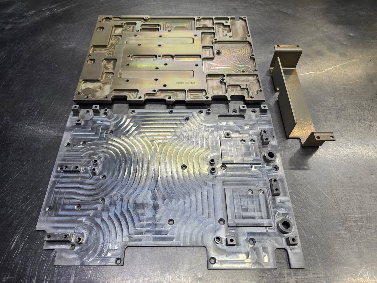 Two machined metal plates with intricate surface patterns and a separate metal bracket placed on a metal work surface.