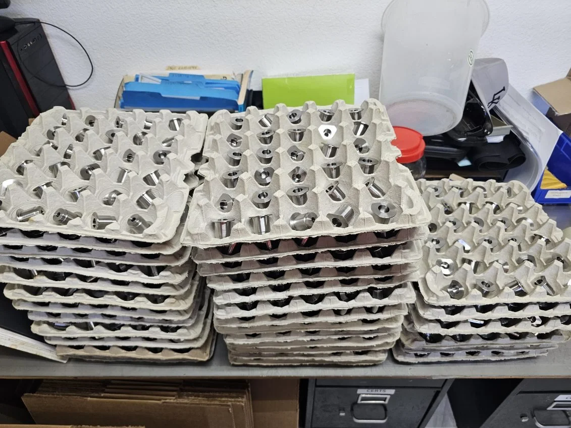 Multiple trays of machined metal engine components on a cluttered workbench.