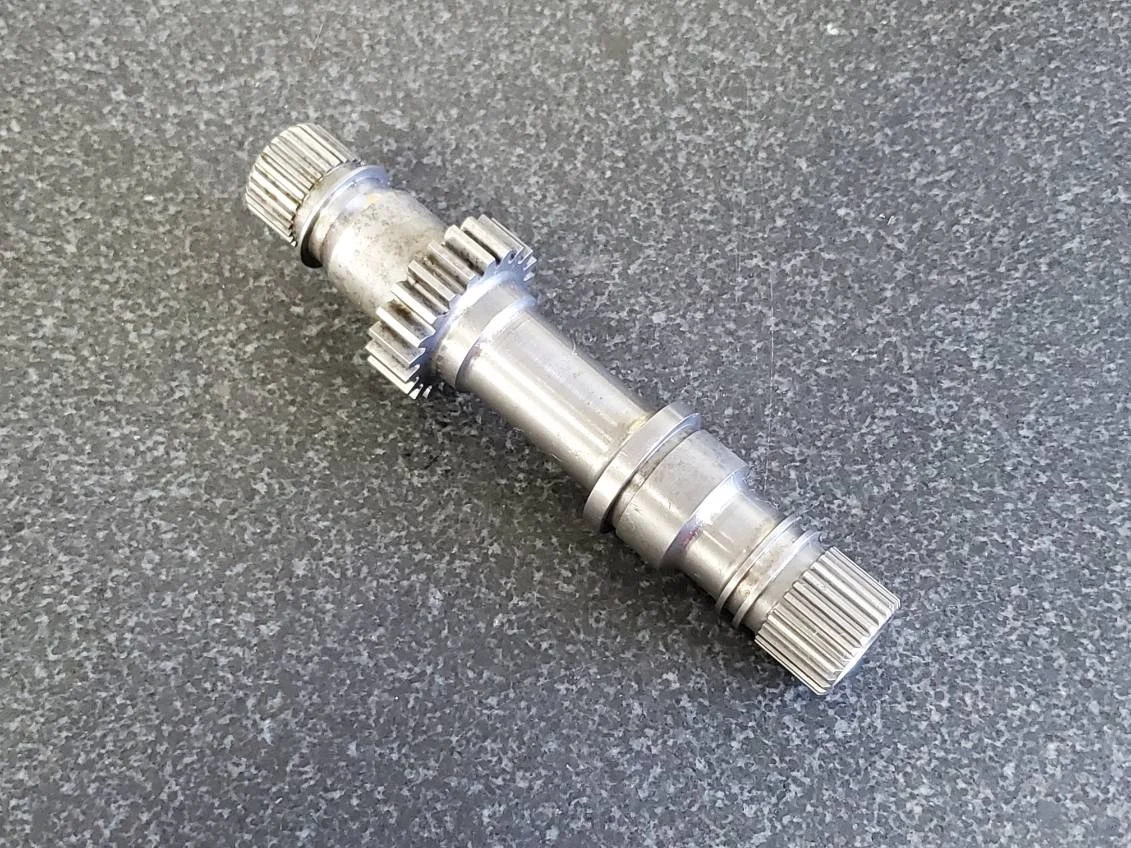 Metal gear shaft with splined ends and gear teeth on a dark textured surface.