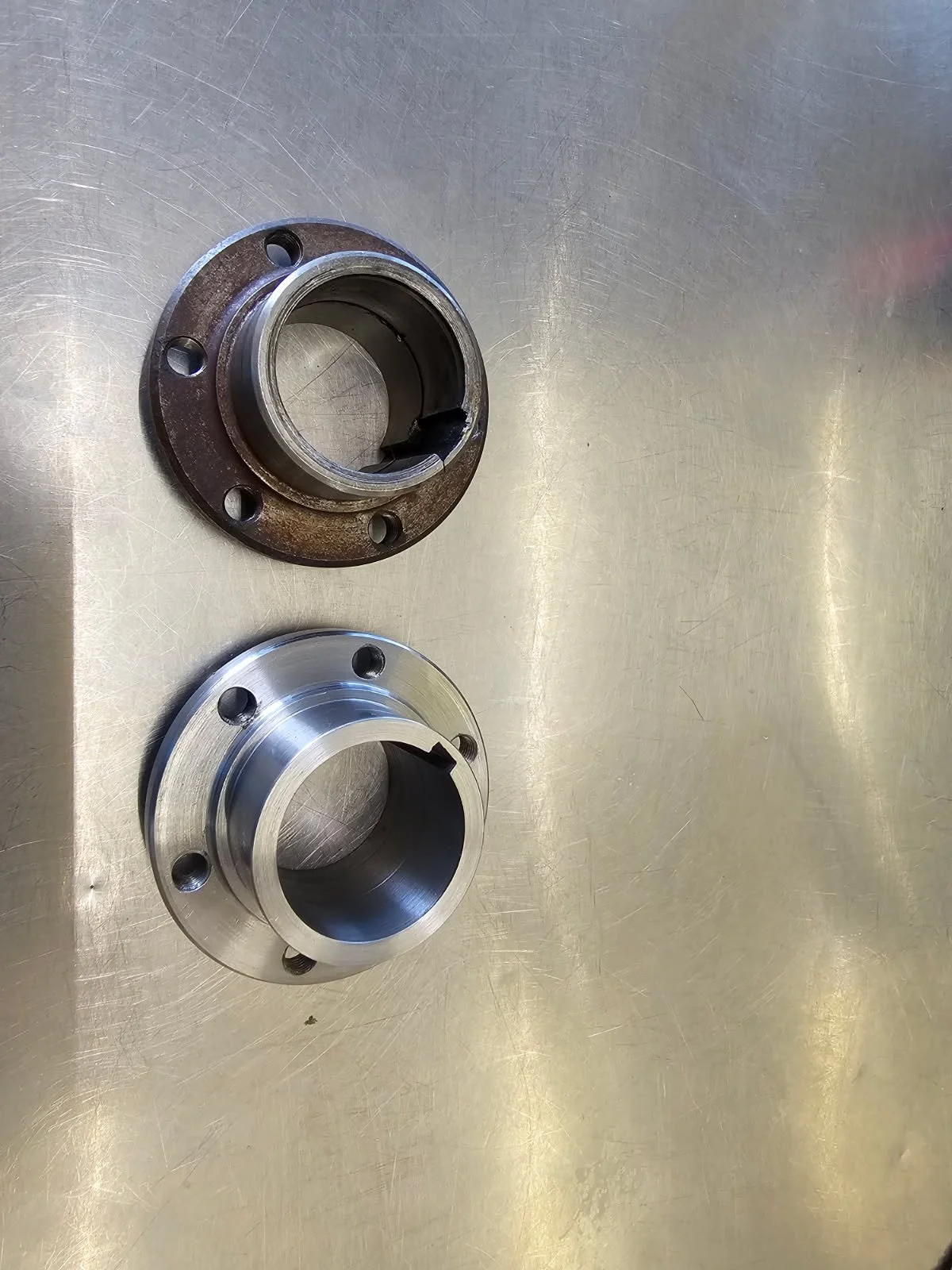 Two metal flange fittings on a metallic work surface, one rusty and one clean, likely used for pipe connections.
