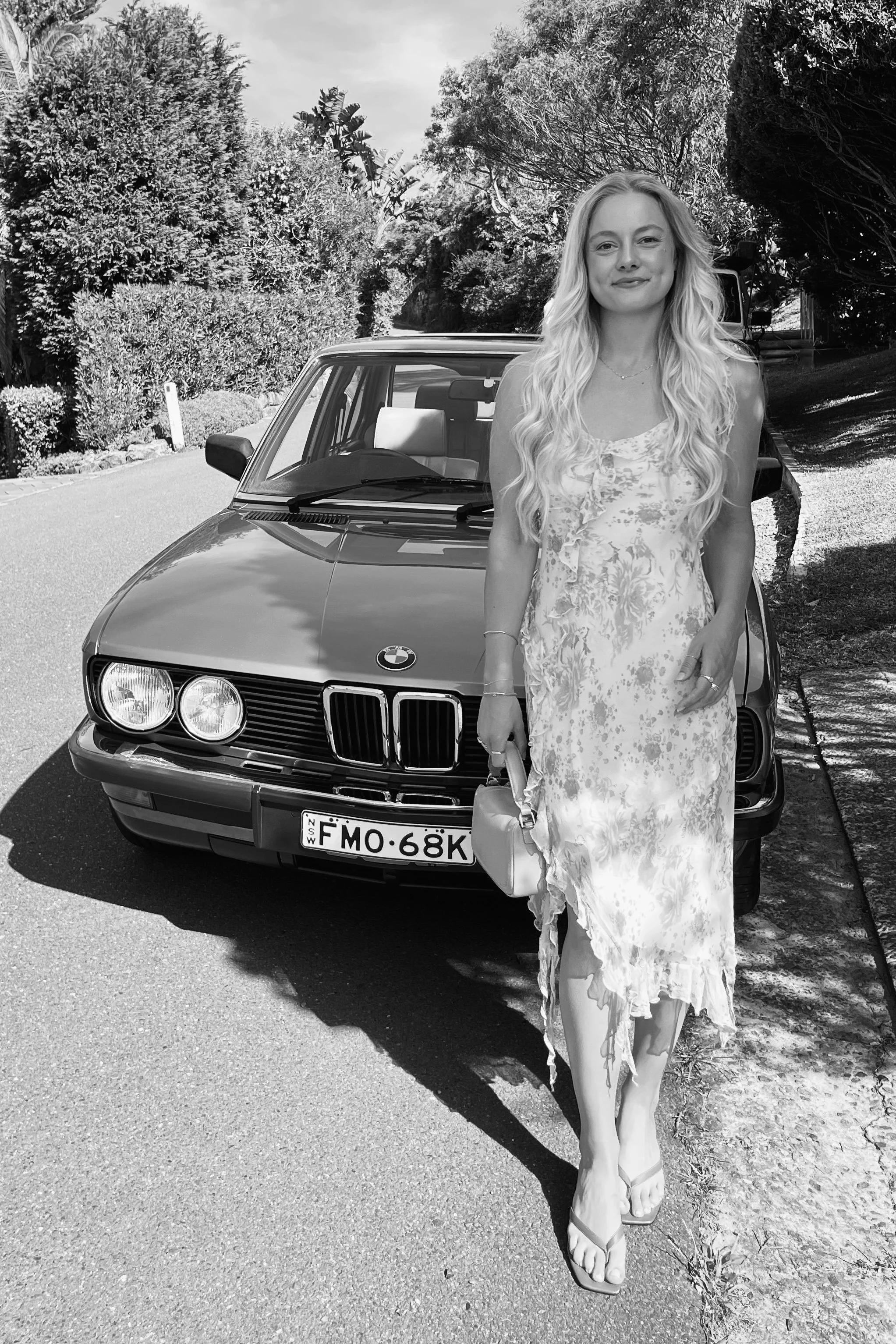 A woman in a floral dress standing in front of a parked car on a road, surrounded by trees and bushes, holding a handbag, with shadows cast on the ground.