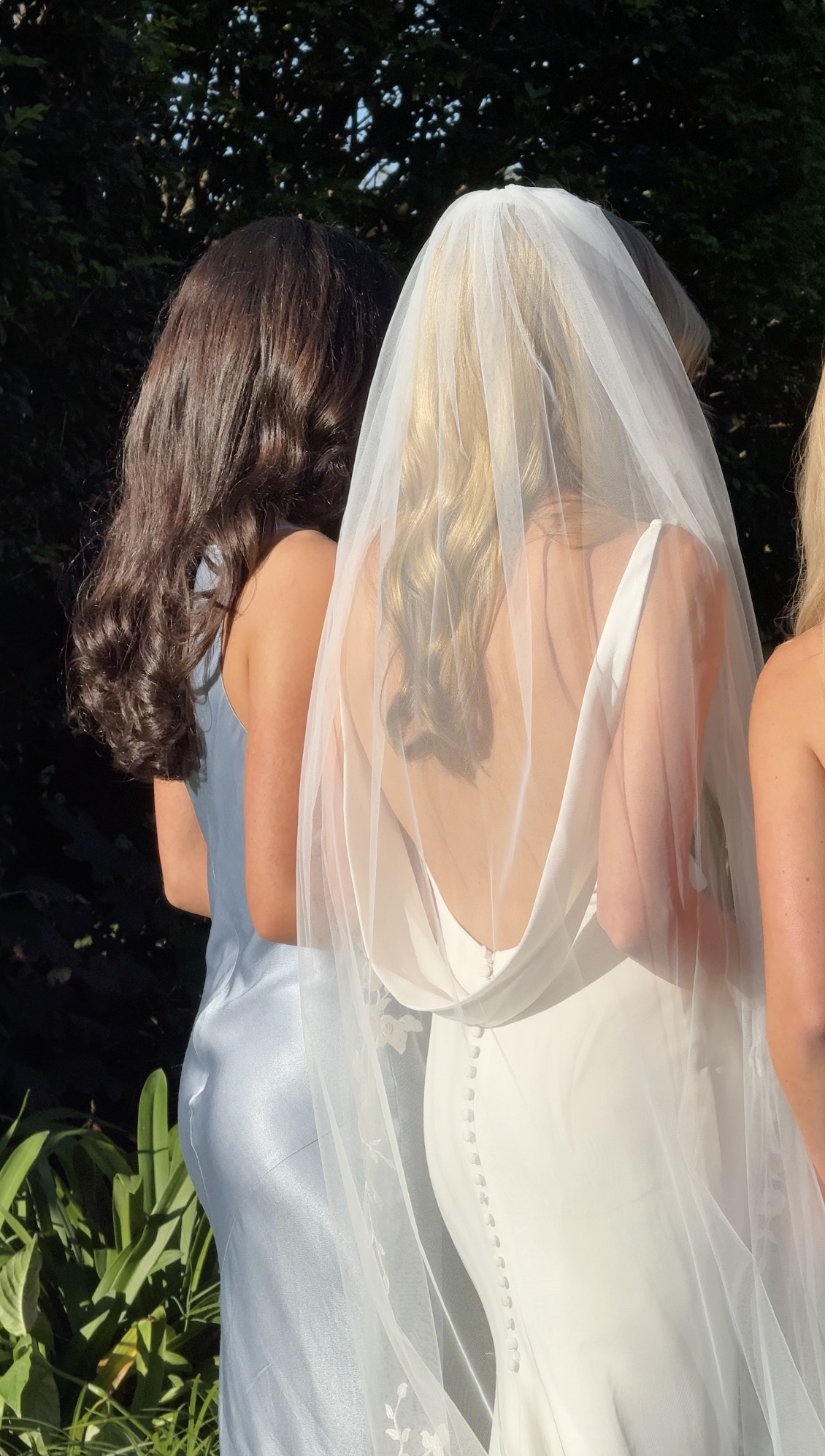 Three women standing outdoors, one wearing a white wedding dress with a veil, and the other two in satin dresses, with a background of green foliage.