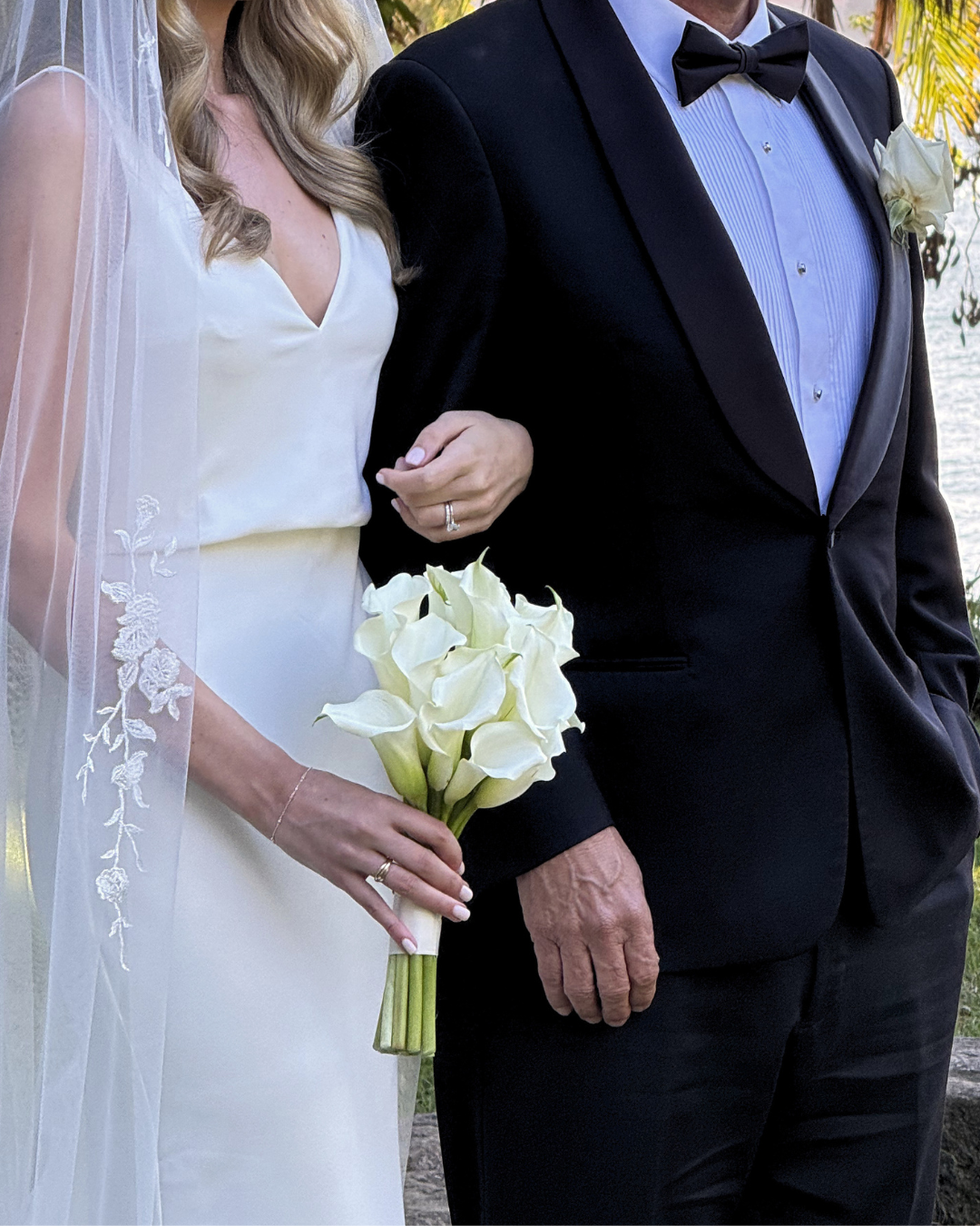 Bride and groom standing outdoors, bride holding a bouquet of white calla lilies, bride wearing a white wedding dress with lace veil, groom in black tuxedo with bow tie, water and foliage in background, during a wedding ceremony.