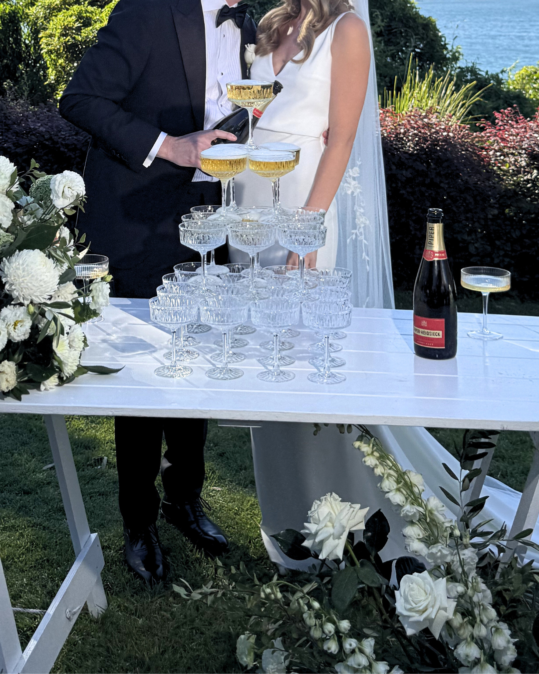 A newlywed couple in formal wedding attire standing behind a table with a champagne tower, decorated with flowers, outdoors near water.