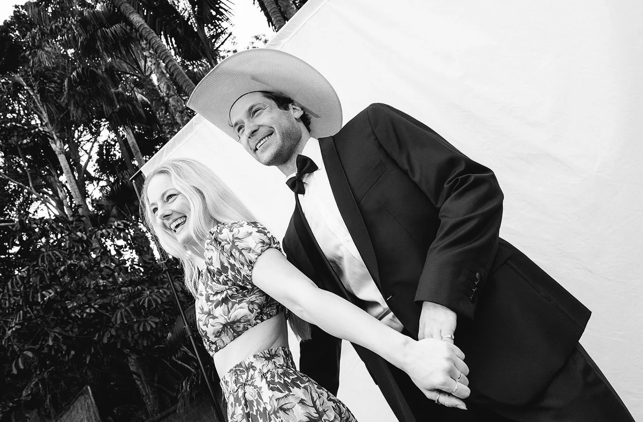 A man and woman holding hands and smiling, with the man wearing a black suit, white shirt, and a large hat, and the woman wearing a floral dress, standing outdoors with trees in the background.