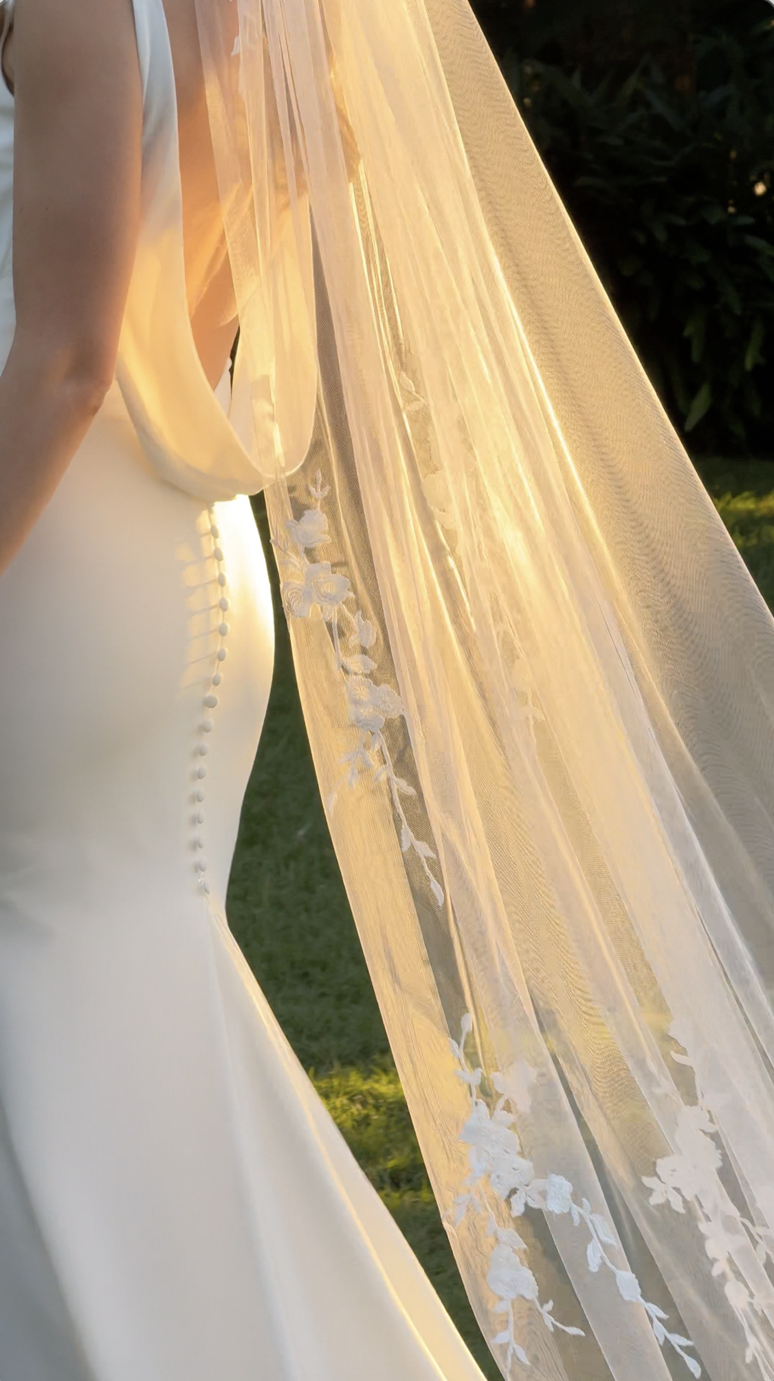 Close-up of a wedding dress with lace details and a sheer veil, bathed in warm sunlight outdoors.