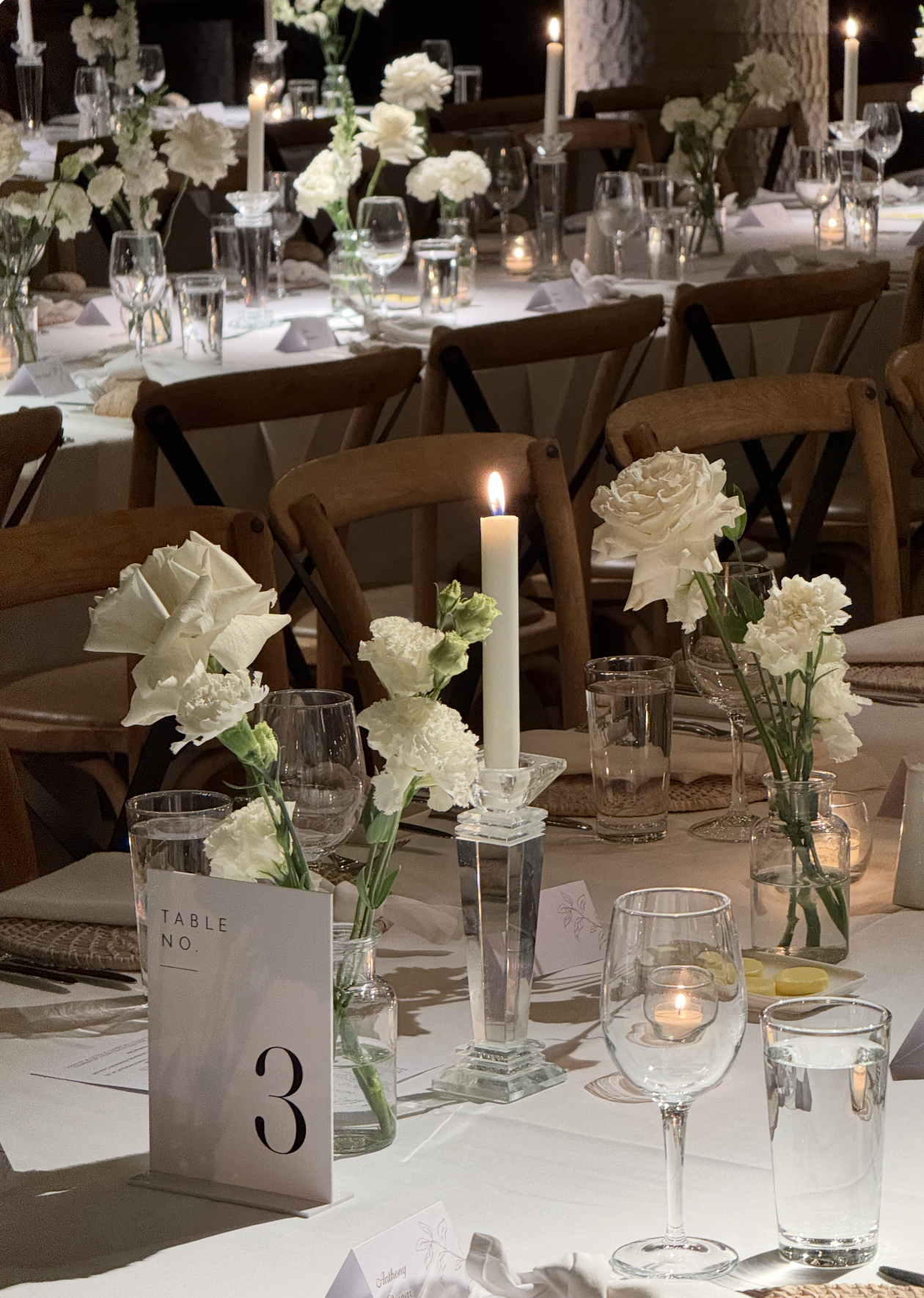 Elegantly decorated banquet table with white flowers in glass vases, tall candles in candelabras, and glassware, set for a formal event or celebration.