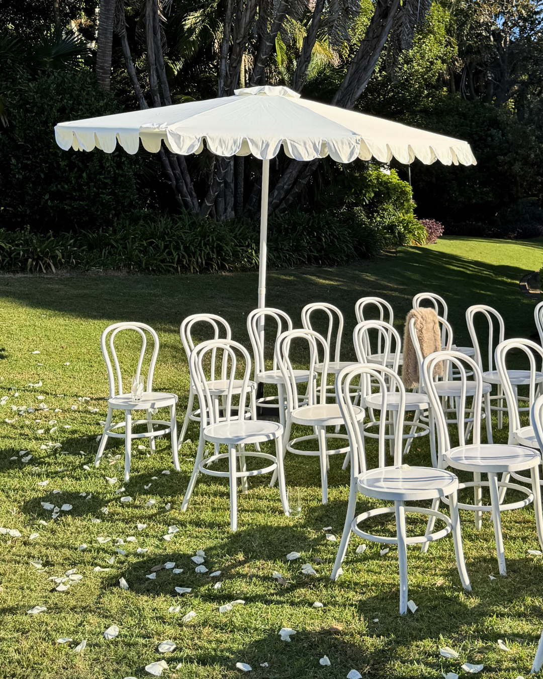 Outdoor wedding or event setup with white chairs arranged in a semi-circle under a large white patio umbrella on a grassy lawn, with flower petals scattered on the ground and lush green trees and bushes in the background.