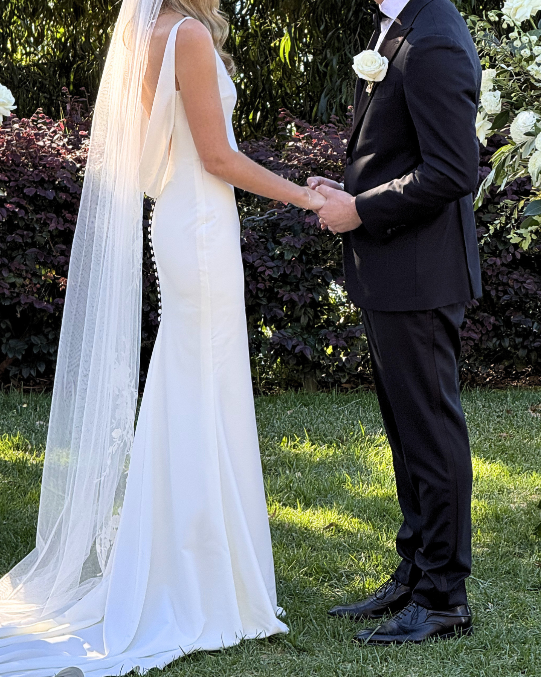 A bride and groom holding hands during a wedding ceremony outdoors, with the bride in a white gown and veil and the groom in a black suit with a white rose boutonniere.