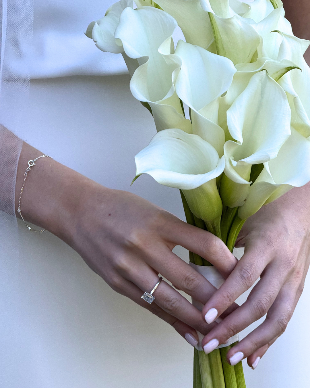 A person's hands holding a bouquet of white calla lilies, wearing a silver ring and a delicate bracelet.