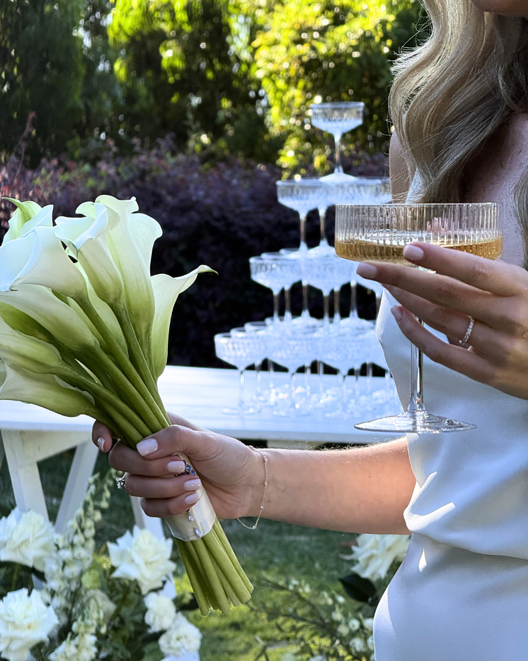 A woman holding a bouquet of white calla lilies and a champagne glass at an outdoor celebration, with a pyramid of empty champagne glasses in the background.