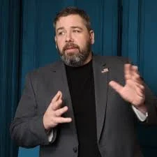 A man with a beard wearing a dark suit jacket and black shirt, standing in front of a blue curtain, gesturing with his right hand as if speaking.