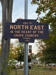 A street sign that reads 'North East in the heart of the Grape Country' in Erie County 16, founded 1794, with trees and a park in the background.