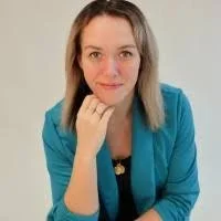 A woman with shoulder-length brown hair wearing a teal blazer, smiling slightly, resting her chin on her hand, against a neutral background.