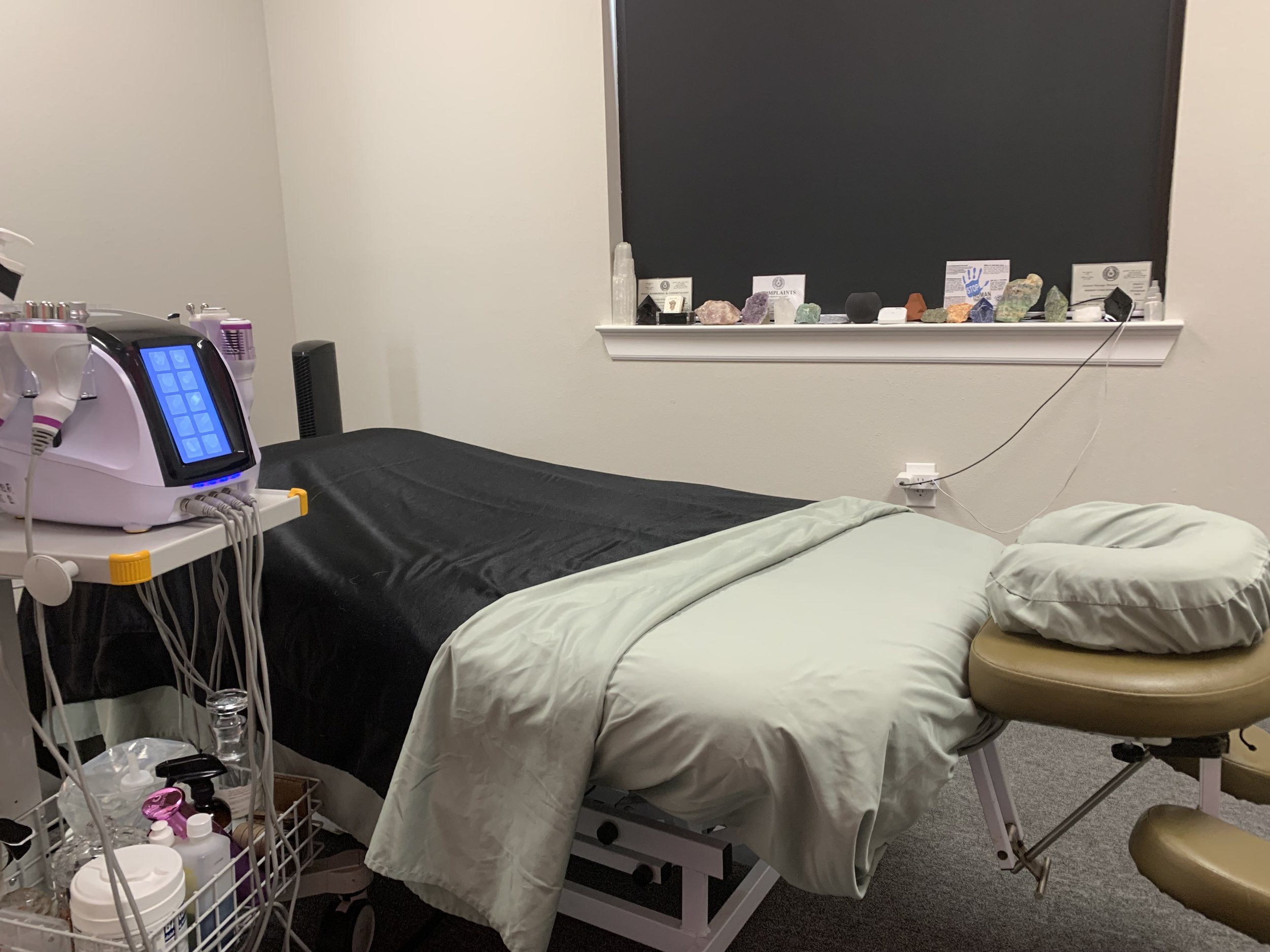 A medical examination room with an examination bed, a medical device with a touchscreen, a stool, and a windowsill with various rocks and certifications.