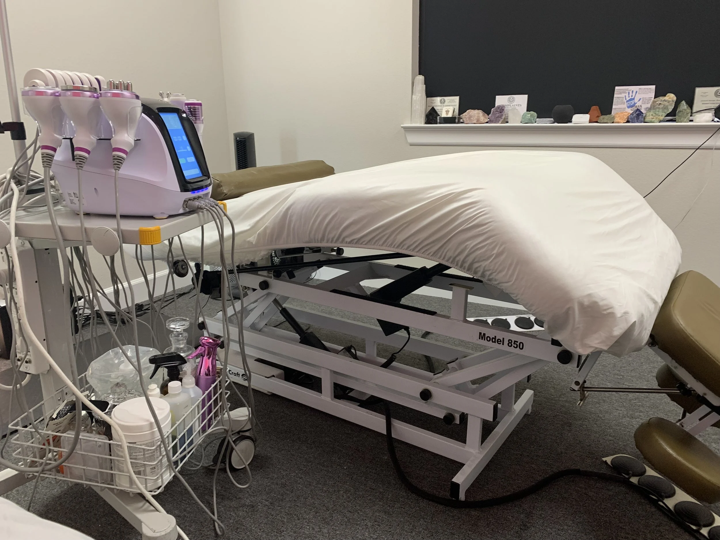 Medical examination room with a hospital bed, various medical devices and supplies, including a machine with multiple probes, a tray with bottles, and a collection of crystals and stones on the windowsill.