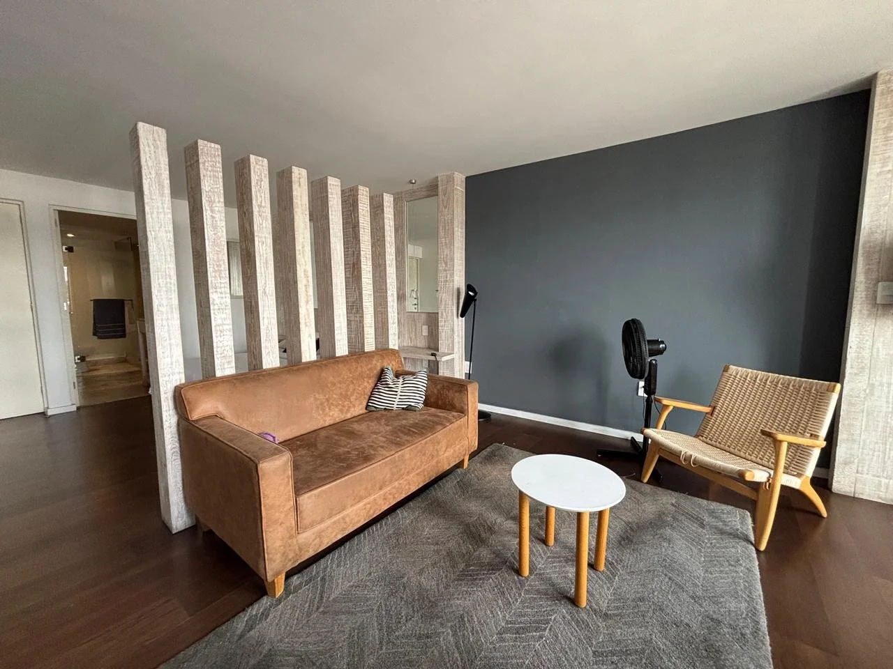 Sala de estar con sillón de terciopelo marrón y silla de madera, mesa blanca pequeña, pared gris oscuro, y división decorativa de madera.