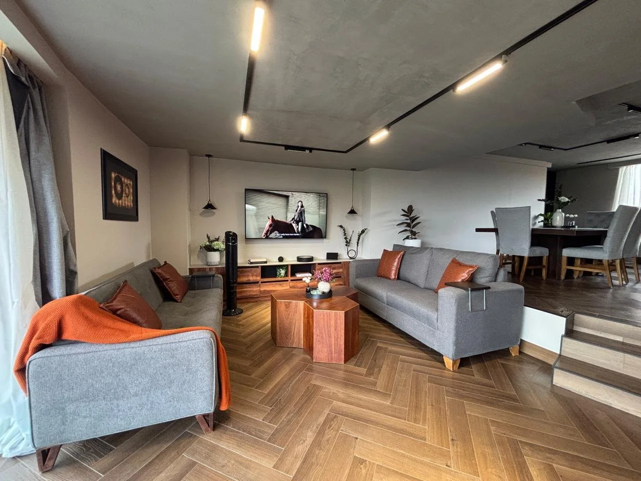 Sala de estar con dos sofás grises, mesa de madera central, televisión en la pared, plantas decorativas y techos con iluminación moderna.