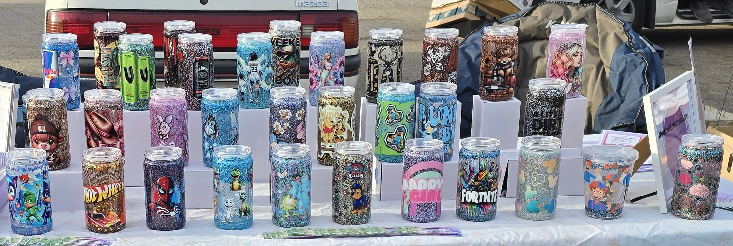 Collection of decorated tumblers with various colorful designs displayed on a table at an outdoor market.