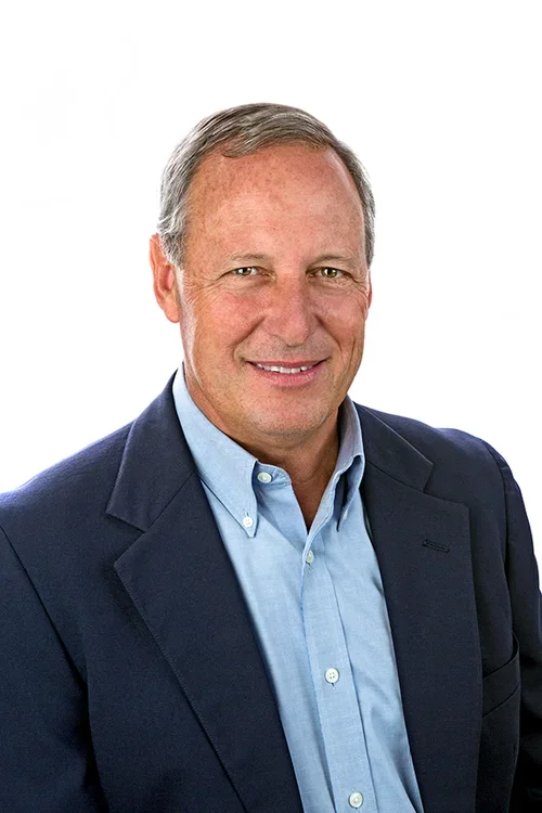 A middle-aged man with short gray hair smiling, wearing a navy suit jacket over a light blue dress shirt, against a white background.
