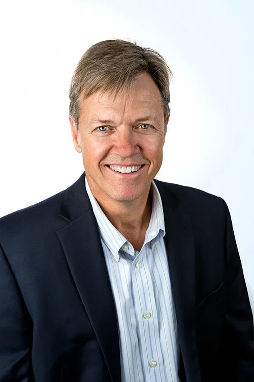 A middle-aged man with light brown hair, smiling, wearing a dark blazer and a striped white shirt, against a plain light background.