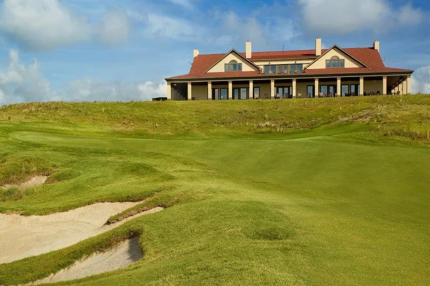 A large house on a hill overlooking a golf course with sand traps, under a partly cloudy sky.
