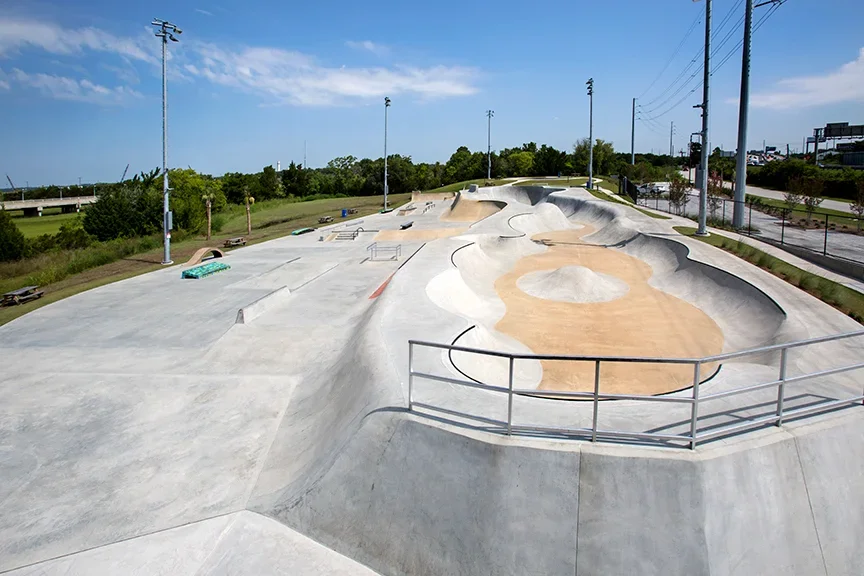An empty outdoor skatepark with concrete ramps, bowls, and rails, surrounded by grass and trees, under a clear blue sky.