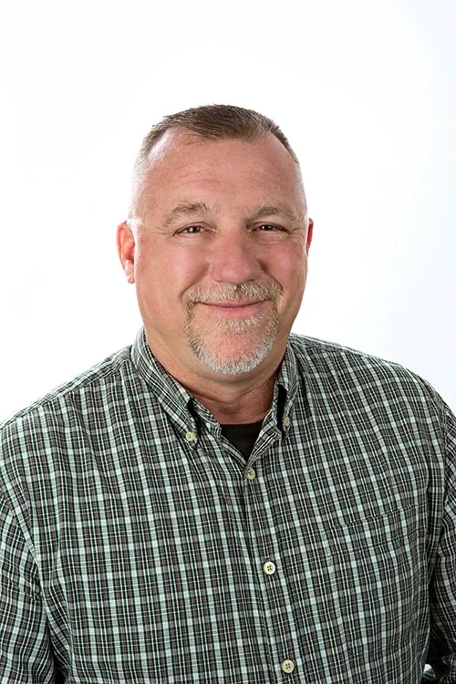 A middle-aged man with short gray hair, a goatee, and a mustache smiling while wearing a green plaid shirt against a white background.