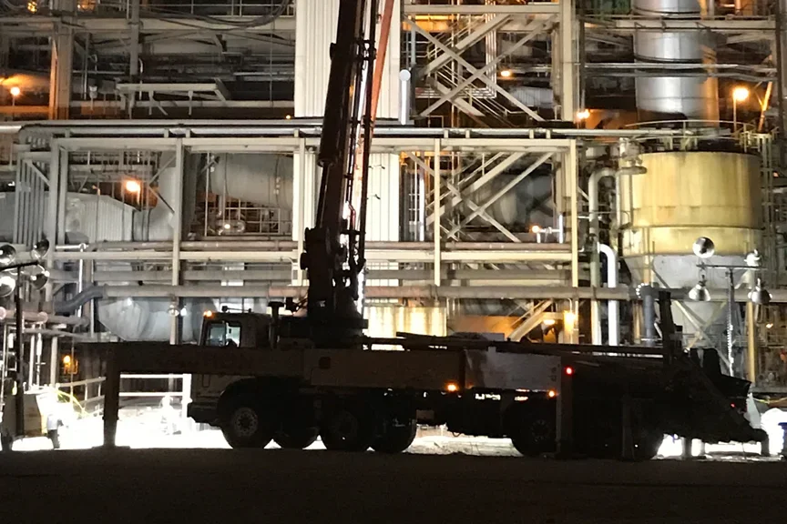 A crane truck at a large industrial facility with extensive pipes and metal structures, illuminated during nighttime.