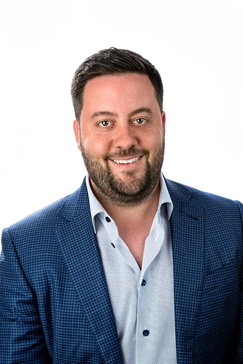Professional headshot of a smiling man with dark hair, beard, wearing a blue checkered blazer and light blue shirt against a white background.