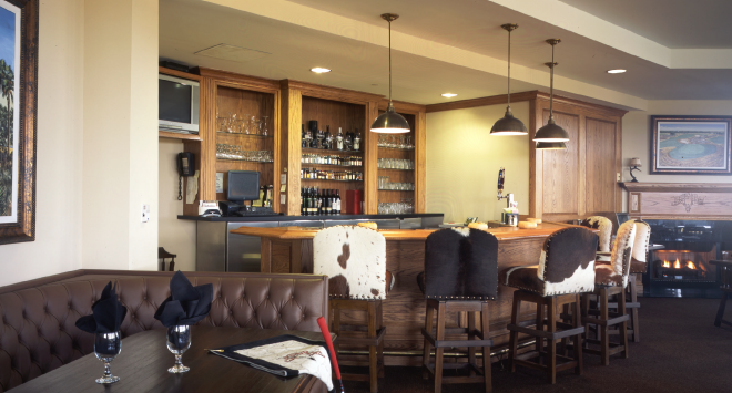 Bar area with wooden shelves stocked with liquor bottles, pendant lights, and bar stools, with a seating area that includes a dining table with glasses and napkins. Fireplace in the background.