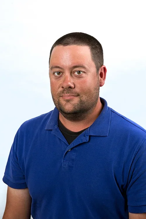 A man with short dark hair and a beard, wearing a blue collared shirt, looking at the camera against a plain white background.
