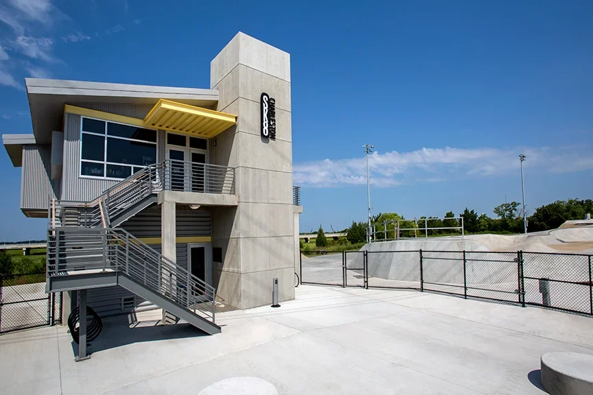 Modern sports facility with a tall concrete building, metal staircase, and a skatepark with ramps, surrounded by a chain-link fence.