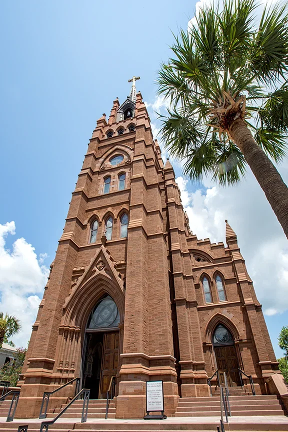 A tall red brick church with Gothic architecture, pointed arches, and stained glass windows, next to a tall palm tree under a blue sky with some clouds.