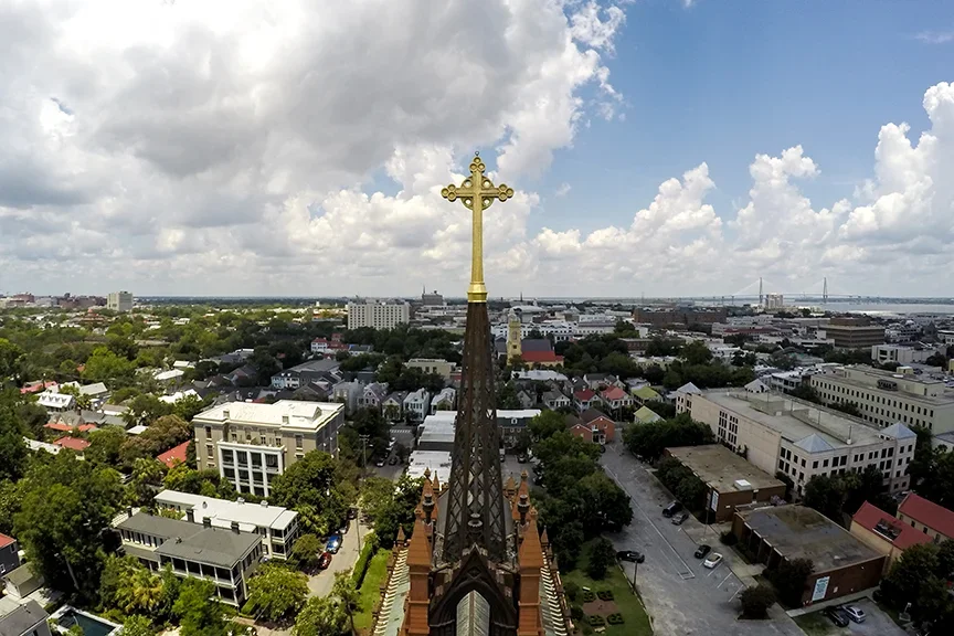 An aerial view of a cityscape featuring a church steeple with a gold cross at the top, surrounded by buildings, trees, and streets under a partly cloudy sky.