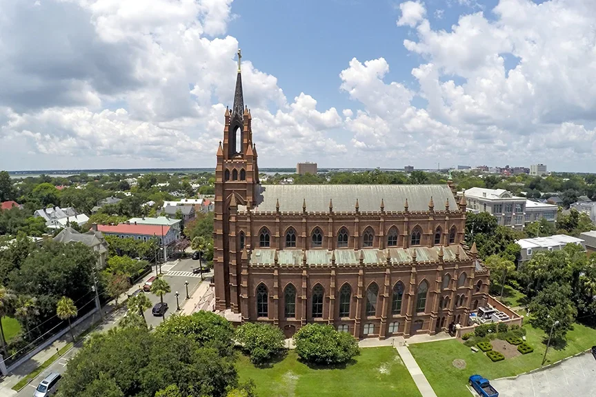 A large, historic church with pointed arch windows and a tall steeple, surrounded by trees and cityscape under a partly cloudy sky.