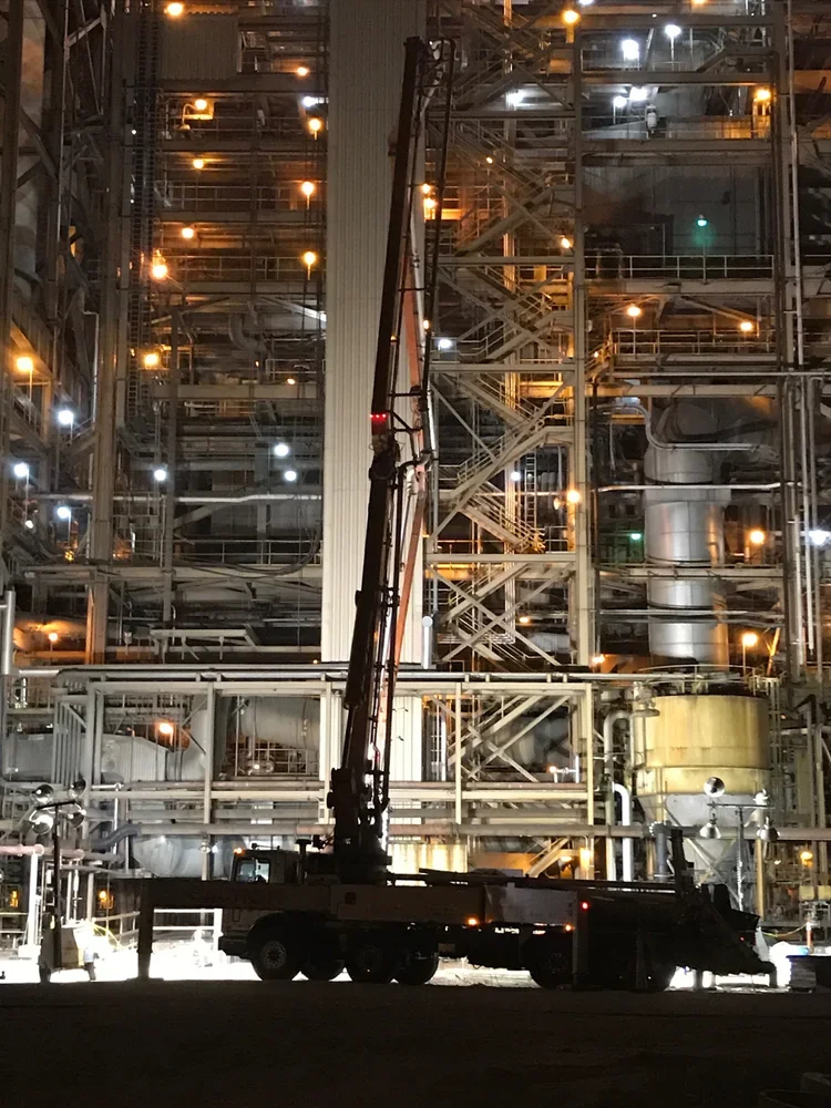 Nighttime view of an industrial facility with numerous pipes, metal structures, and bright lights. A large crane is positioned in the foreground.