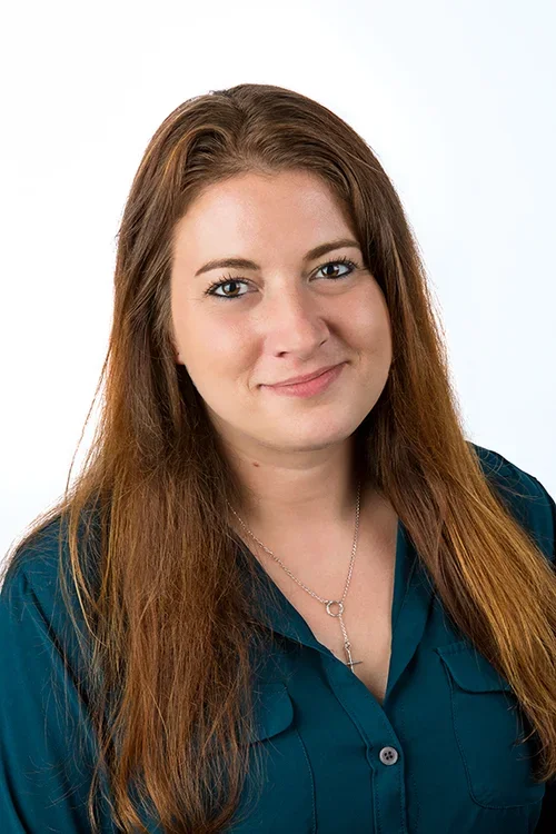 A woman with long brown hair smiling, wearing a dark teal blouse and a silver necklace, against a plain white background.