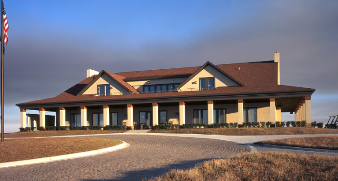 Large beige house with a red-tiled roof, white columns, and multiple windows, situated on a grassy landscape with a curved paved driveway and an American flag on a flagpole.