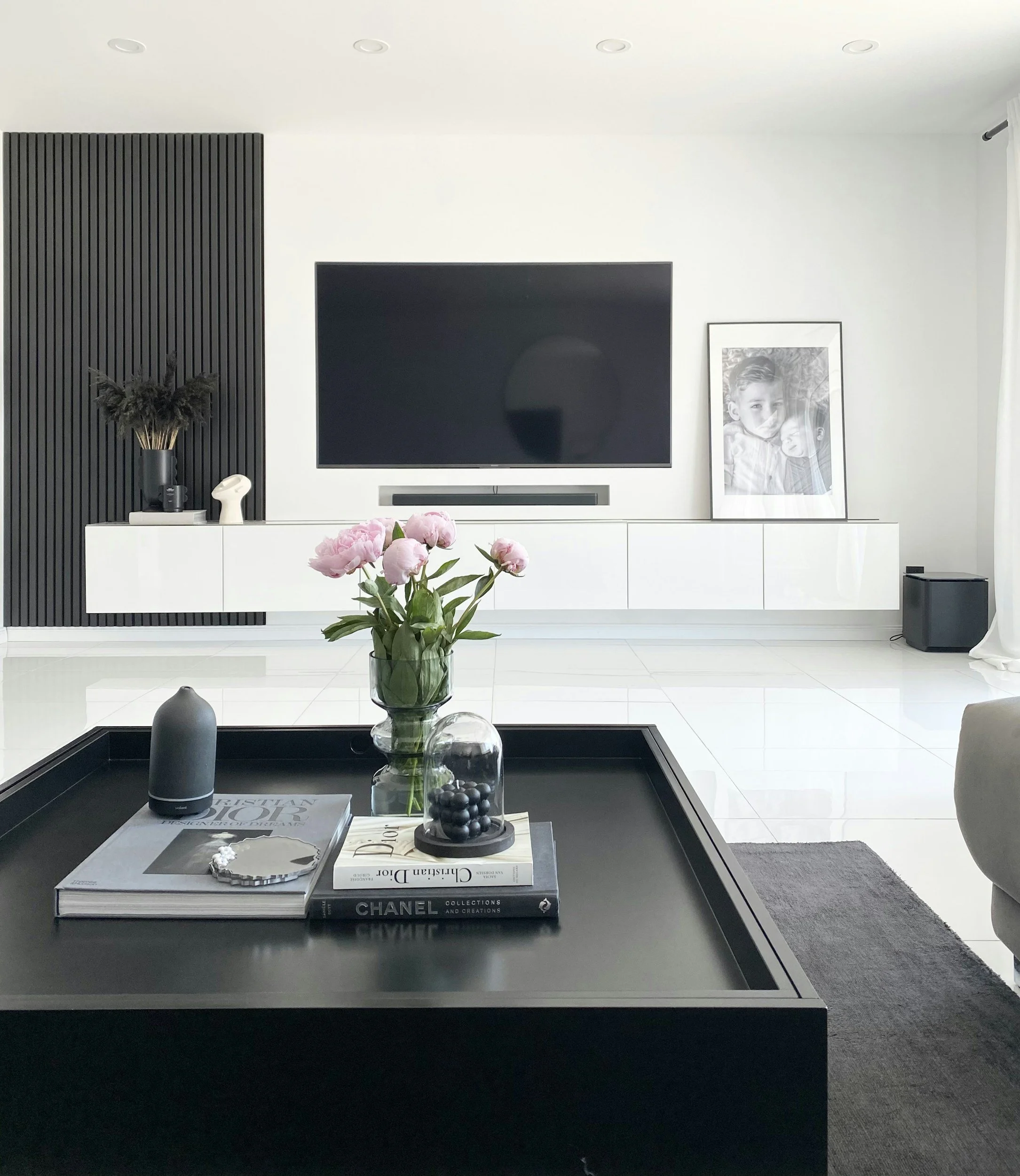 Modern living room with television, framed black-and-white photo of children, black accent wall, vase with pink flowers, books, and decorative objects on a black coffee table, white walls, and curtains.