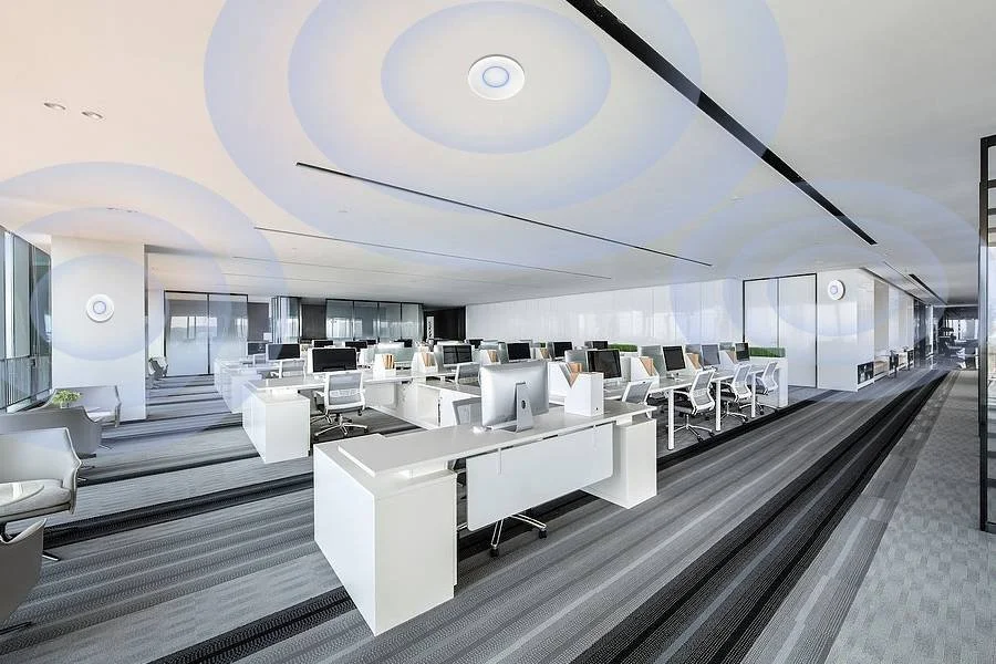 Modern office space with white desks, ergonomic chairs, desktop computers, and large windows in a high-rise building. Image portrays professionally installed PoE Wi-Fi access points providing seamless coverage for a high-traffic network.