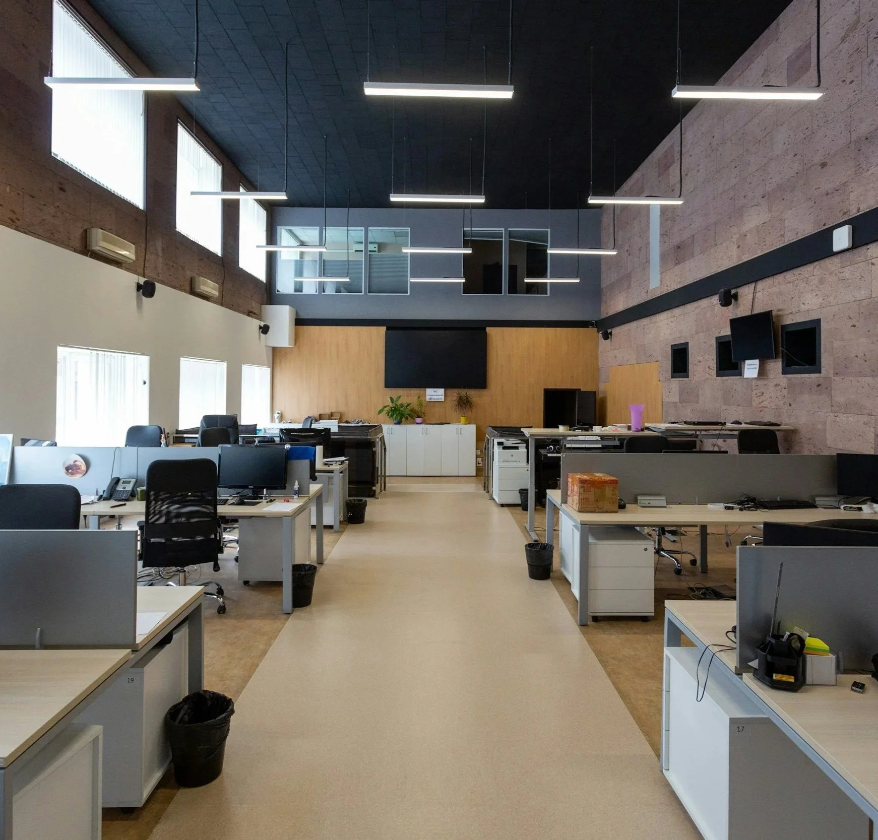 An open-plan modern office with multiple desks, black chairs, and computer equipment. The room features a high ceiling with suspended fluorescent lights, large windows, and a wall with a mounted television and decorative plants. Commercial networking