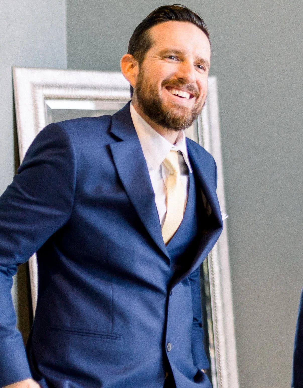 A man with a beard and short dark hair wearing a navy blue suit, white shirt, and light yellow tie smiling and standing indoors in front of a framed mirror.