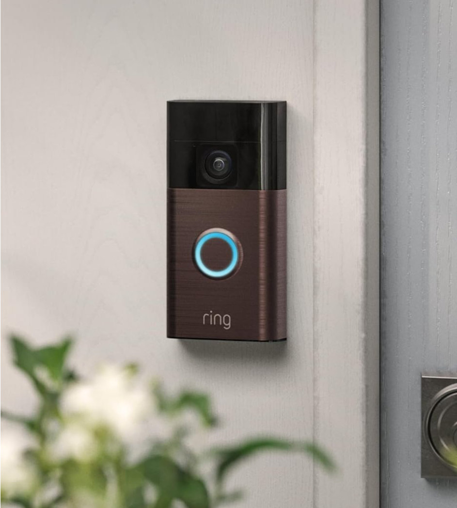 Close-up of a Ring video doorbell mounted on a white exterior wall near a door with a metal hinge.