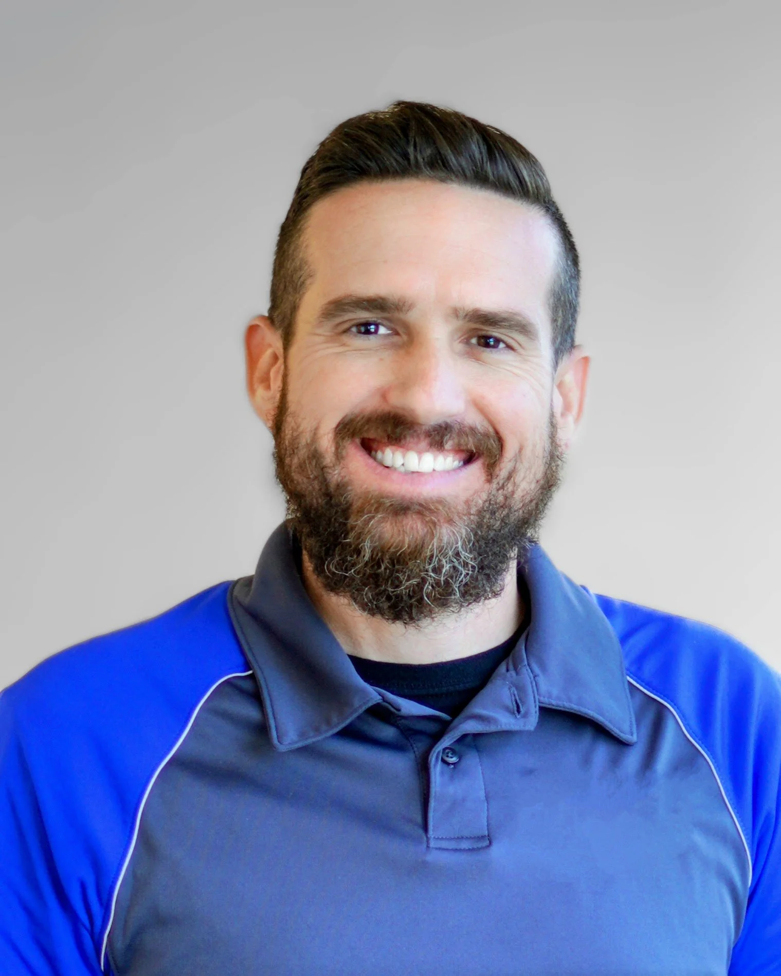 Close-up of a smiling man with dark hair, beard, and mustache, wearing a blue sports jacket and black shirt, standing against a plain light gray background.