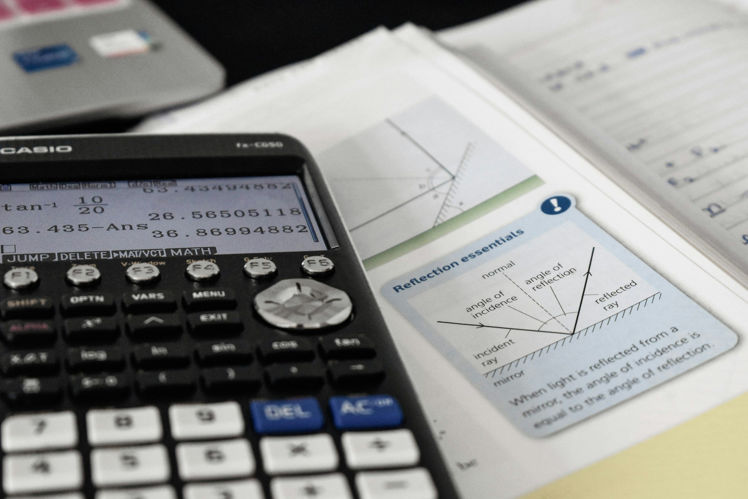 A scientific calculator displaying numbers and equations, resting on an open textbook with diagrams and text about reflection and incident rays. There are also some sheets of paper on the background. Math Support for Nearby Local Tutoring.