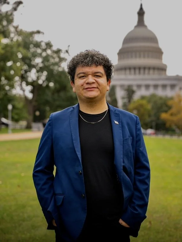 A man with curly hair wearing a navy blazer and black shirt standing outdoors in front of the U.S. Capitol building. Founder of Nerd Alert AZ, Alexis Delgado Rodriguez