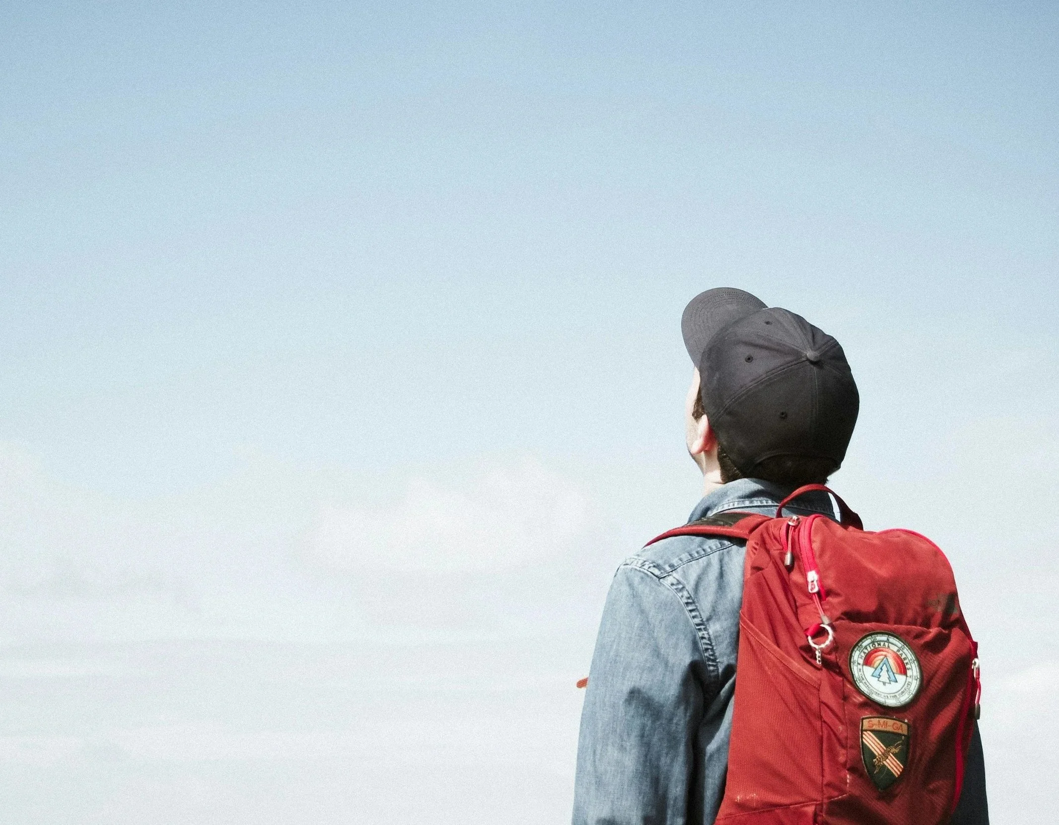Person wearing a black cap, gray jacket, and red backpack standing outdoors against a pale sky. Looking to the horizon of Academic Opportunity